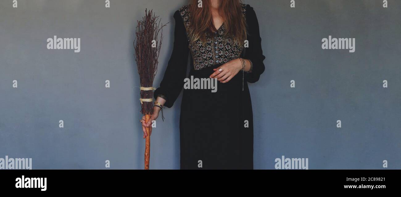 Artificially expanded image. Female woman wiccan witch standing against dark textured blue purple wall, wearing black dress, holding witch besom broom Stock Photo