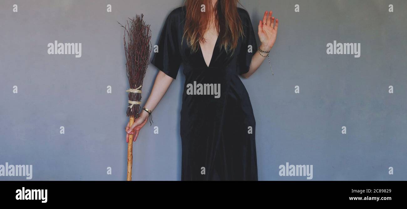 Artificially expanded image. Female woman wiccan witch standing against dark textured blue purple wall, wearing a black dress, holding a witch broom Stock Photo