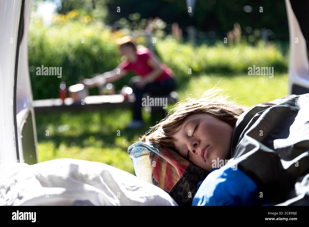 Portrait of a boy sleeping in a van with open doors, mother preparing breakfast outside on a porch in nature Stock Photo