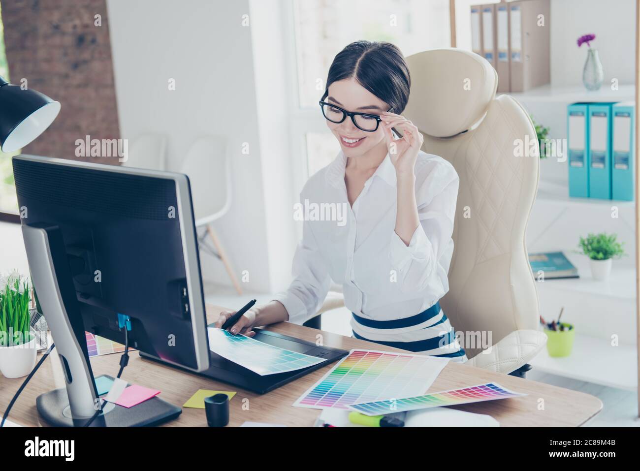 Intelligent young asian graphic designer is working with computer and colors samples, stylus, smiling. She is a successful self employed artist Stock Photo