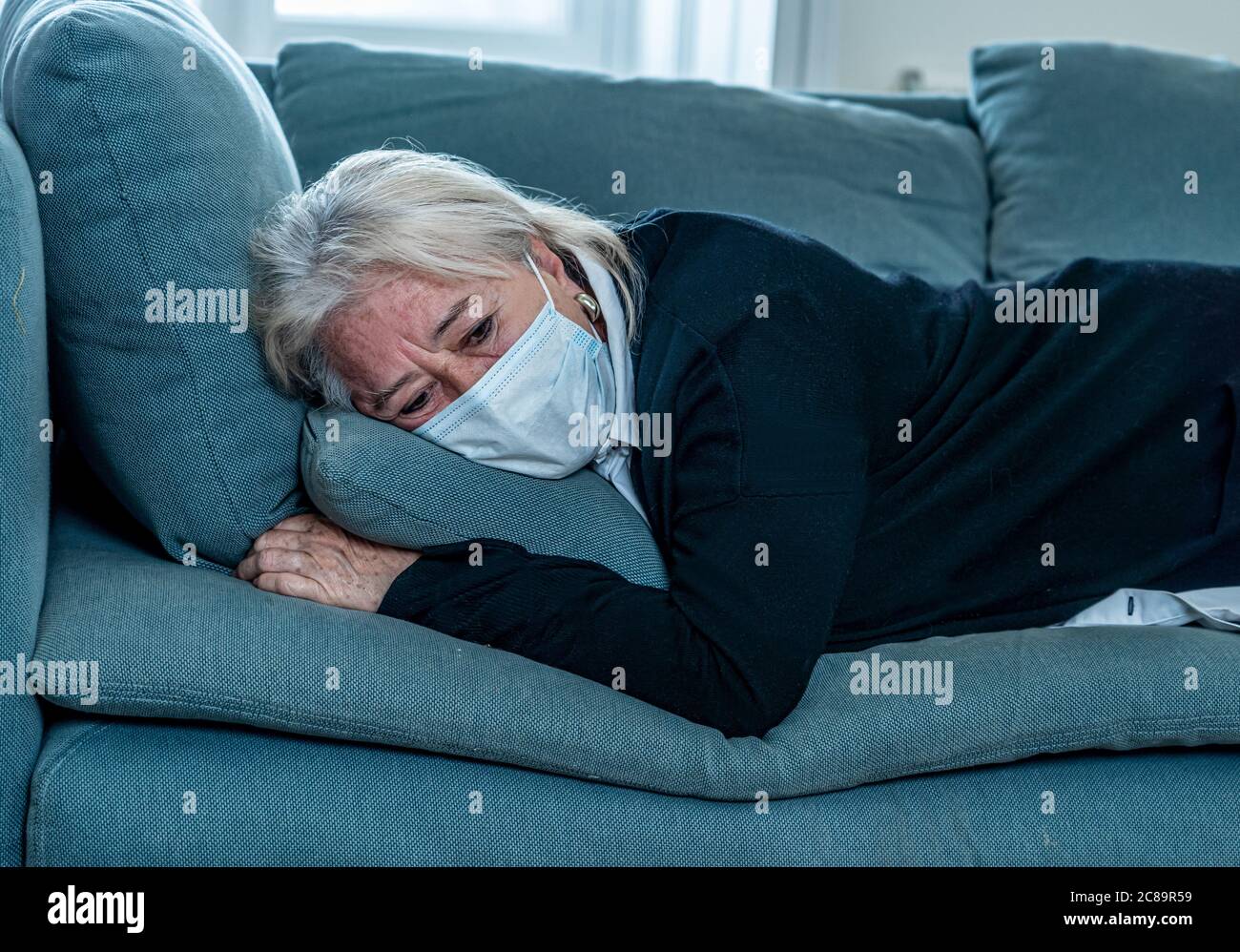 Lonely depressed senior widow woman with protective mask crying on couch isolated at home, sad and worried missing husband and family in COVID-19 deat Stock Photo