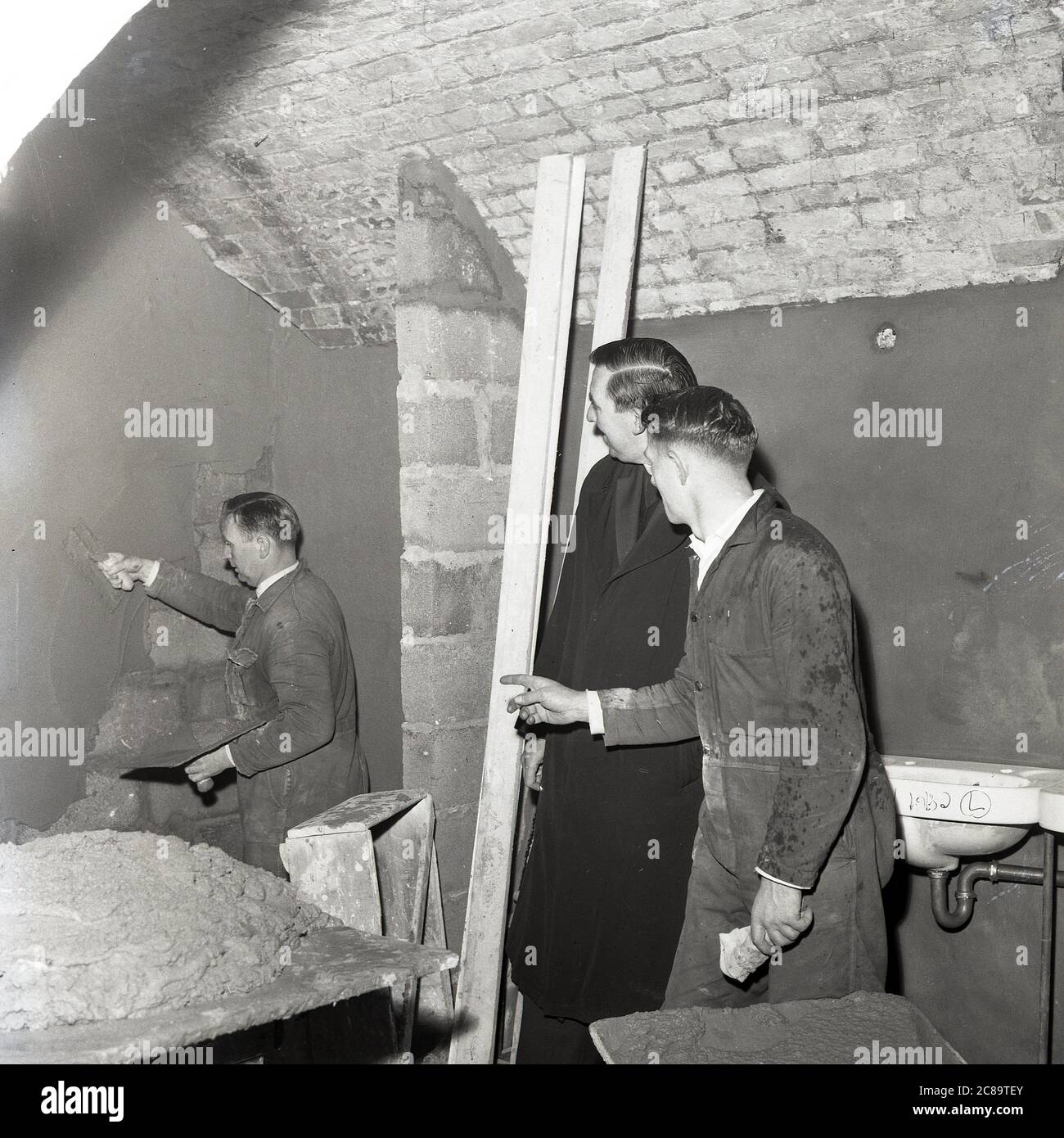 1960s, historical, repairs to the crypt, St. Pauls, Deptford, Southeast London, a worker plastering one of the basements brick wal's. Priest with workmen undertaking repairs  to the crypt. In the 1980s the basement was the venue for raves and gigs including the band, the Stone Roses Stock Photo