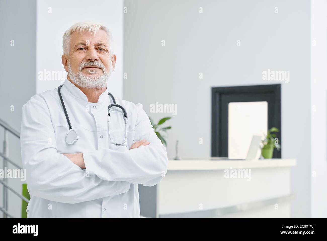 Portrait of eldery caucasian male doctor in white lab coat. Selective focus of professional therapist with stethoscope on neck posing with arms crossed and looking at camera, reception on background. Stock Photo