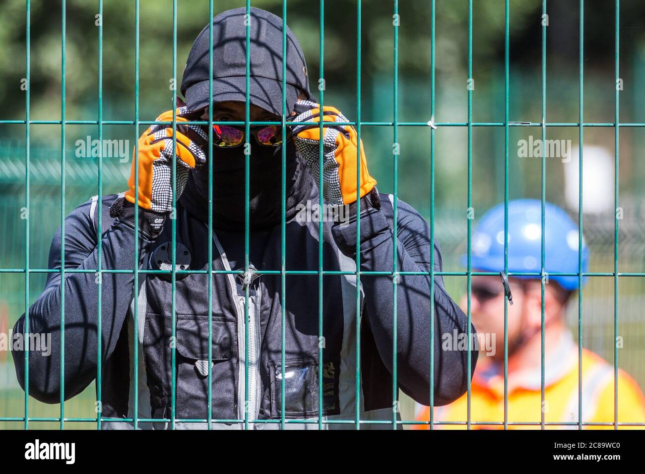 Harefield, UK. 22 July, 2020. A masked HS2 security guard observes a press photographer during works for the HS2 high-speed rail link close to Harvil Road. Environmental activists from HS2 Rebellion and Stop HS2 continue to protest against HS2, which is currently projected to cost £106bn and will remain a net contributor to CO2 emissions during its projected 120-year lifespan, from a series of wildlife protection camps along its route. Credit: Mark Kerrison/Alamy Live News Stock Photo