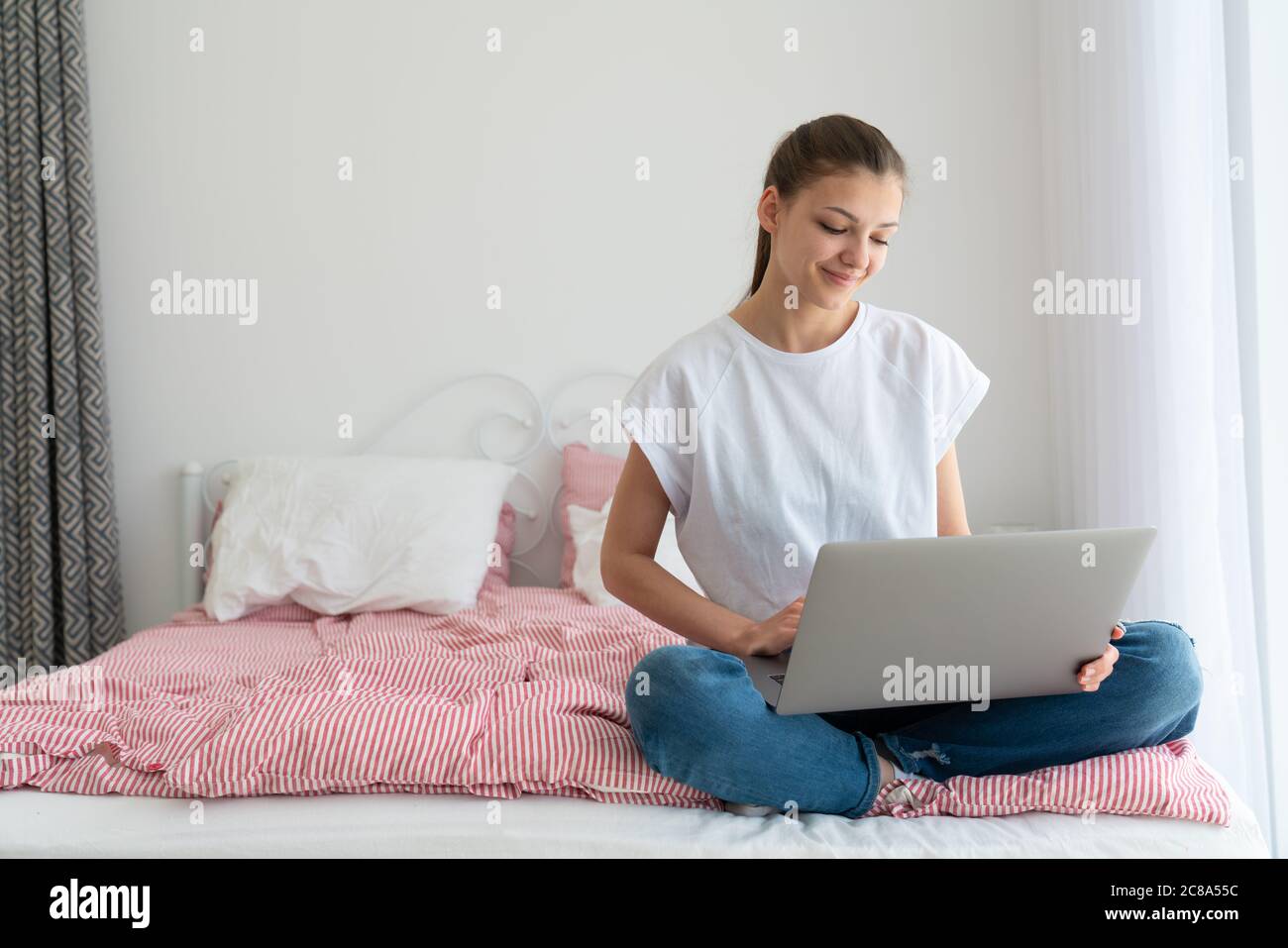 Beautiful girl using laptop and smiling while sitting on bed and studying at home Stock Photo