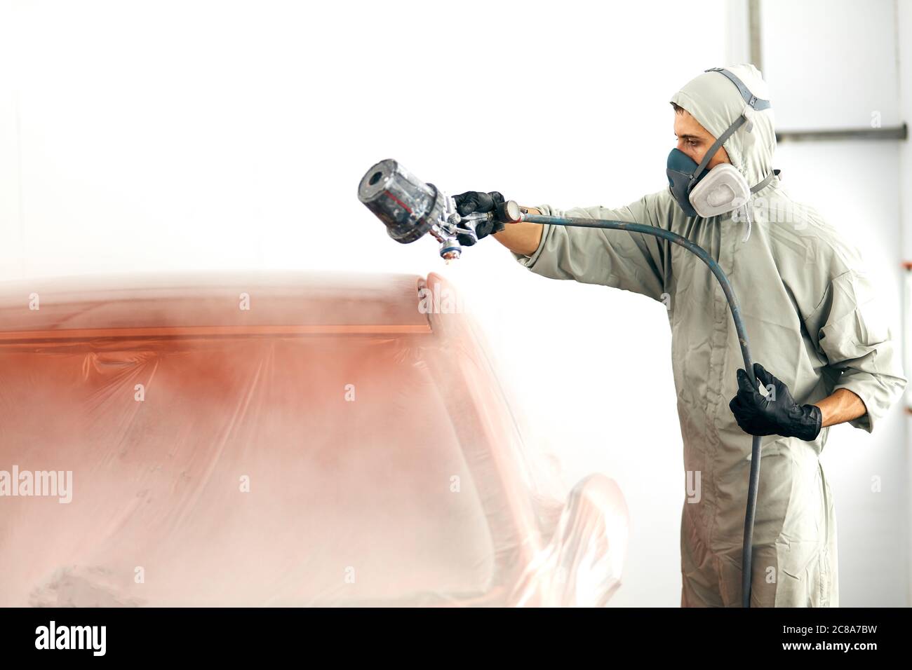 automobile painter in protective workwear and respirator painting car body in paint chamber