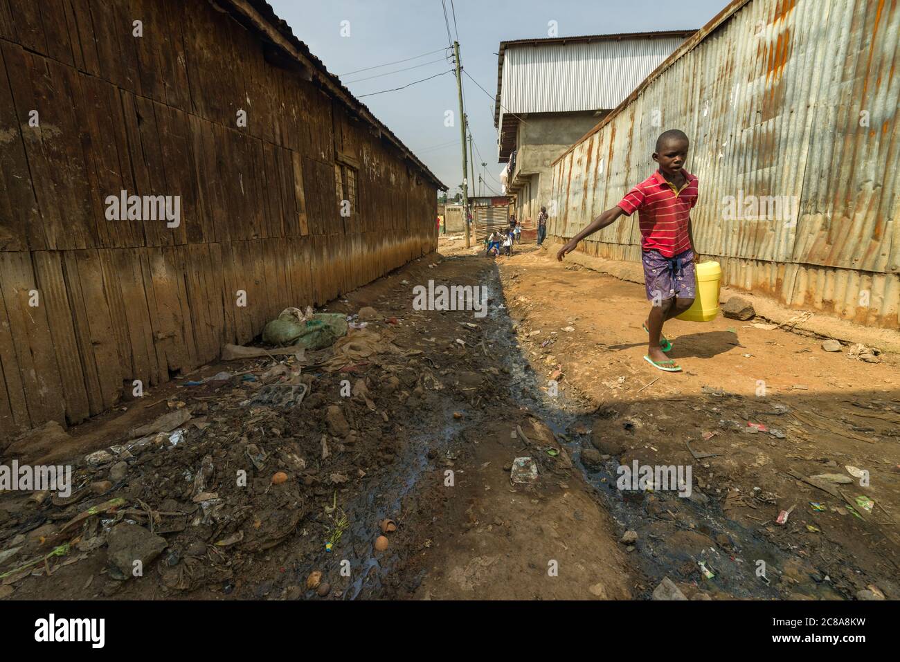A young Kenyan boy carries a water container down a dirt alley past a building with discarded waste along it, Korogocho, Nairobi, Kenya Stock Photo