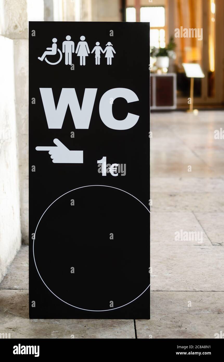 Black signboard in the streets pointing to a nearby public toilet on payment, with a round empty space and a restaurant on background Stock Photo
