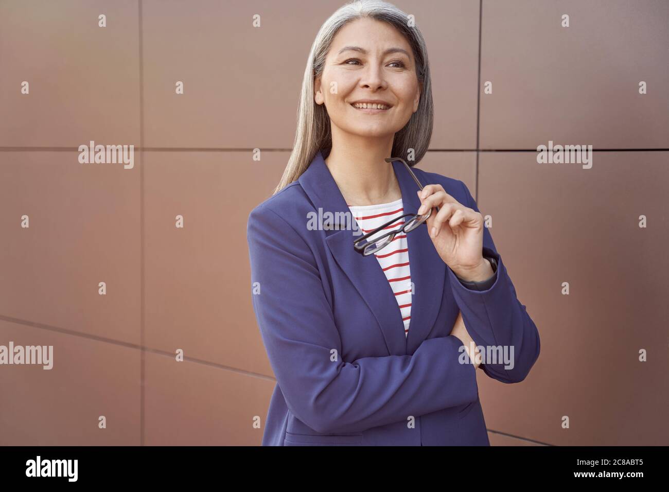Thinking about business. Portrait of a beautiful happy mature business woman in classic wear holding eyeglasses, looking aside and smiling while standing against the wall outdoors. Business people Stock Photo