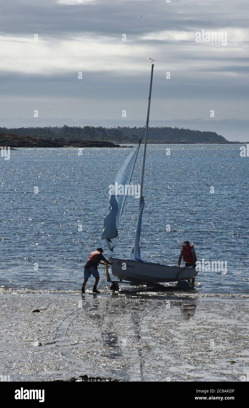 Saanich, British Columbia, Canada 22 July 2020  - Two men prepare to launch an Enterprise,  a Bermuda rigged sailing dinghy with a double-chined hull and distinctive blue sails, from the beach at Cadboro-Gyro Park in to Cadboro Bay on a hot summer day in the greater Victoria municipality of Saanich.  Alamy Live News/Don Denton Stock Photo