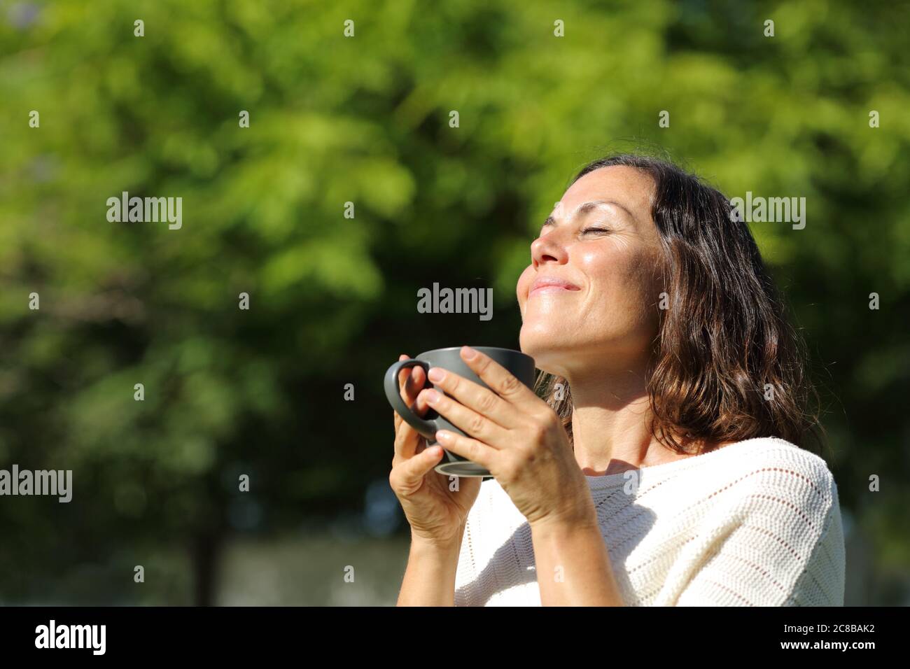 Satisfied adult woman smelling coffee cup standing in a green park at summer Stock Photo
