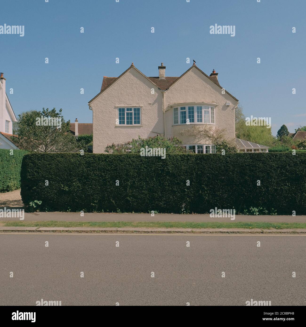 A typical M Shaped / Double Pitched / Double Gabled Roof suburban house half hidden behind a green garden hedge and residential street with blue sky Stock Photo