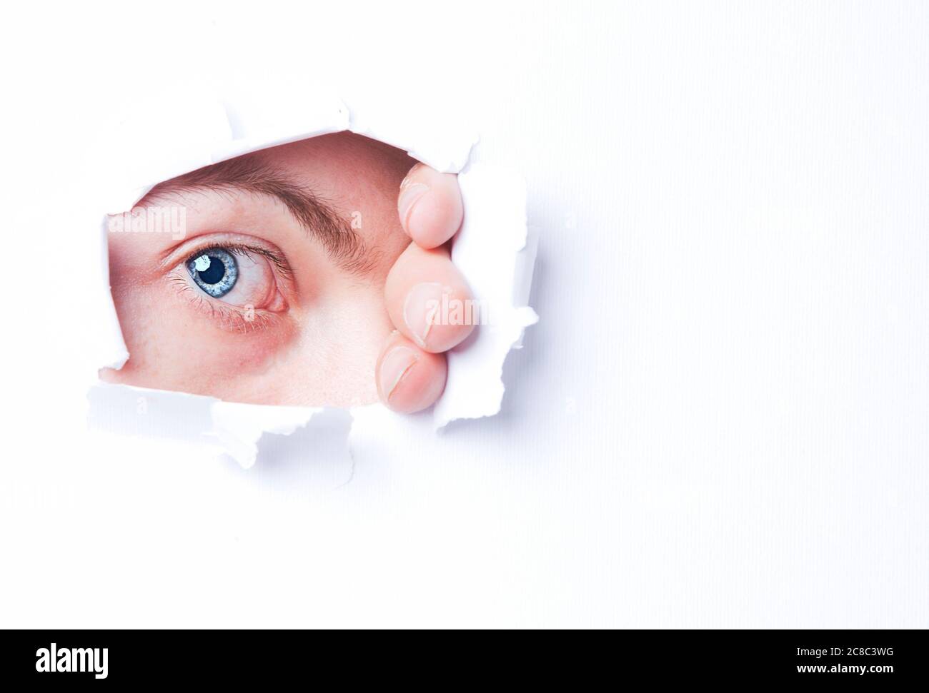 Mans blue eye peering through through ripped paper Stock Photo