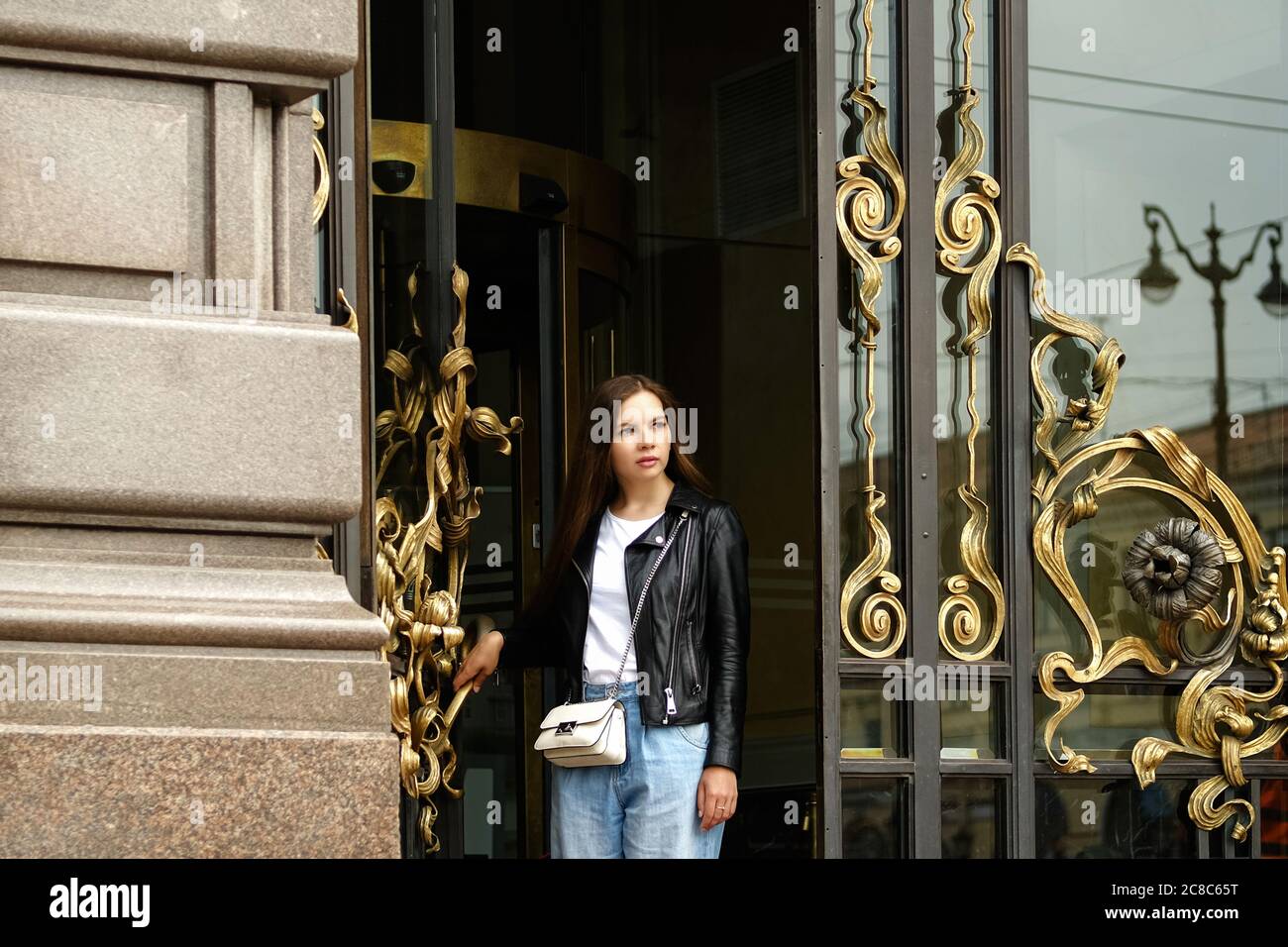 Young woman with long hair going out of old vintage building through ornated glass door to the street of a big city. Saint Petersburg, Russia. Stock Photo