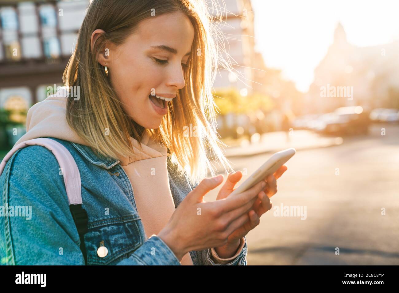 Cheerful young blonde girl using mobile phone while walking on a street ...