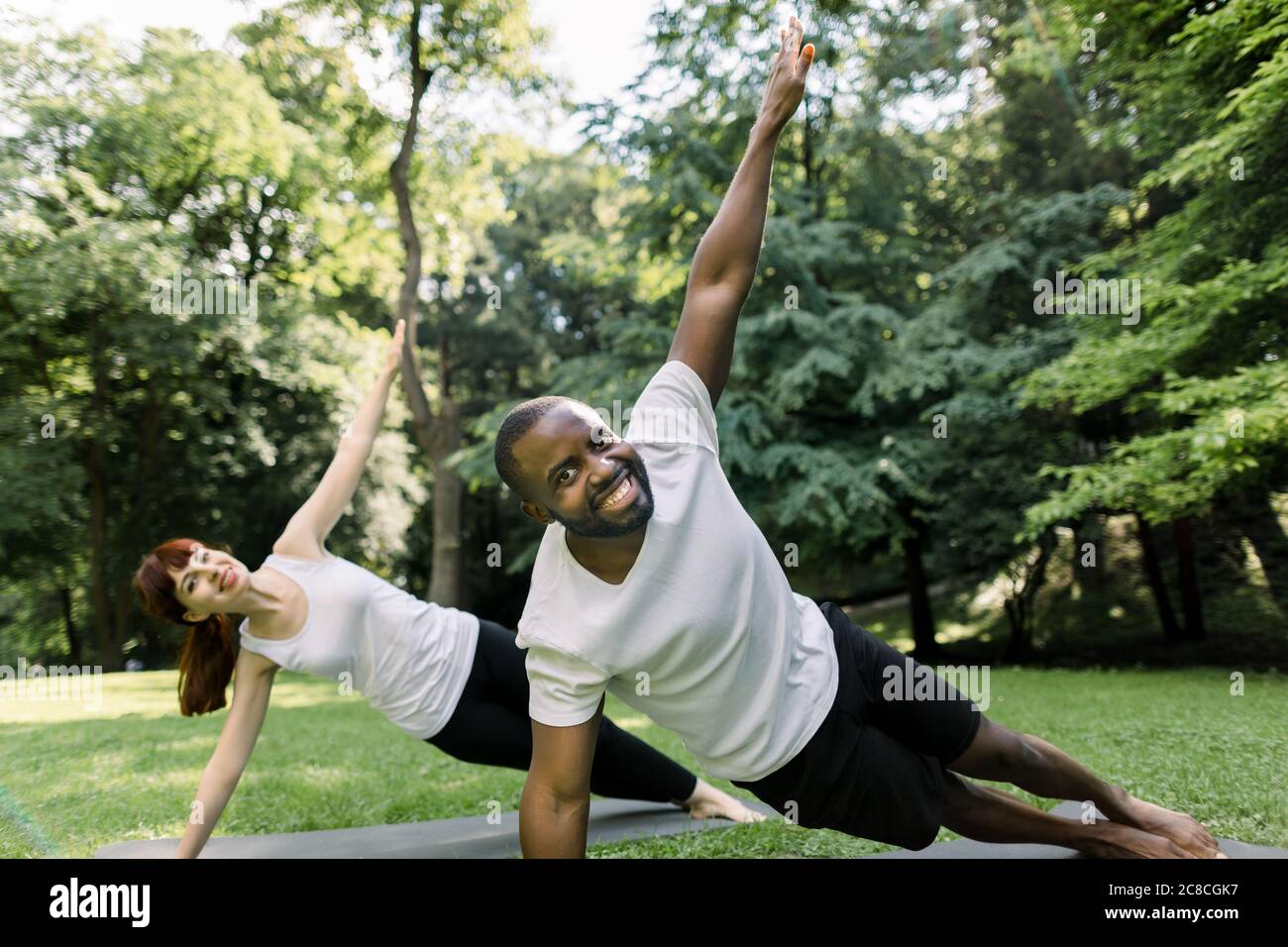 Multiethnic joyful young man and woman exercising in a park, holding side-plank position with one arm raised up. Fit couple training outdoors in the Stock Photo