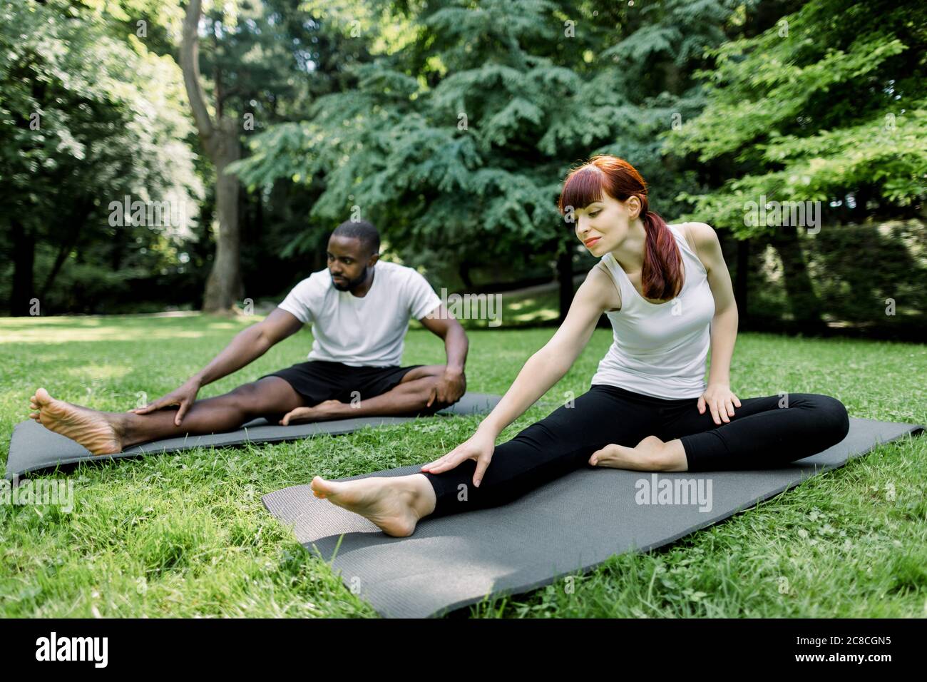Multiethnic couple doing yoga in nature. Young Caucasian woman performs yoga asanas outdoors in a park, together with her handsome African boyfriend Stock Photo