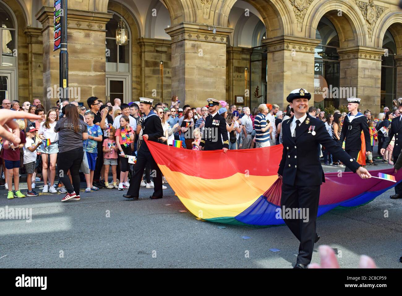 Pride parade passing in front of Radisson Hotel Manchester UK. Royal Navy in uniform carrying giant rainbow pride flag smiling with crowd. Stock Photo