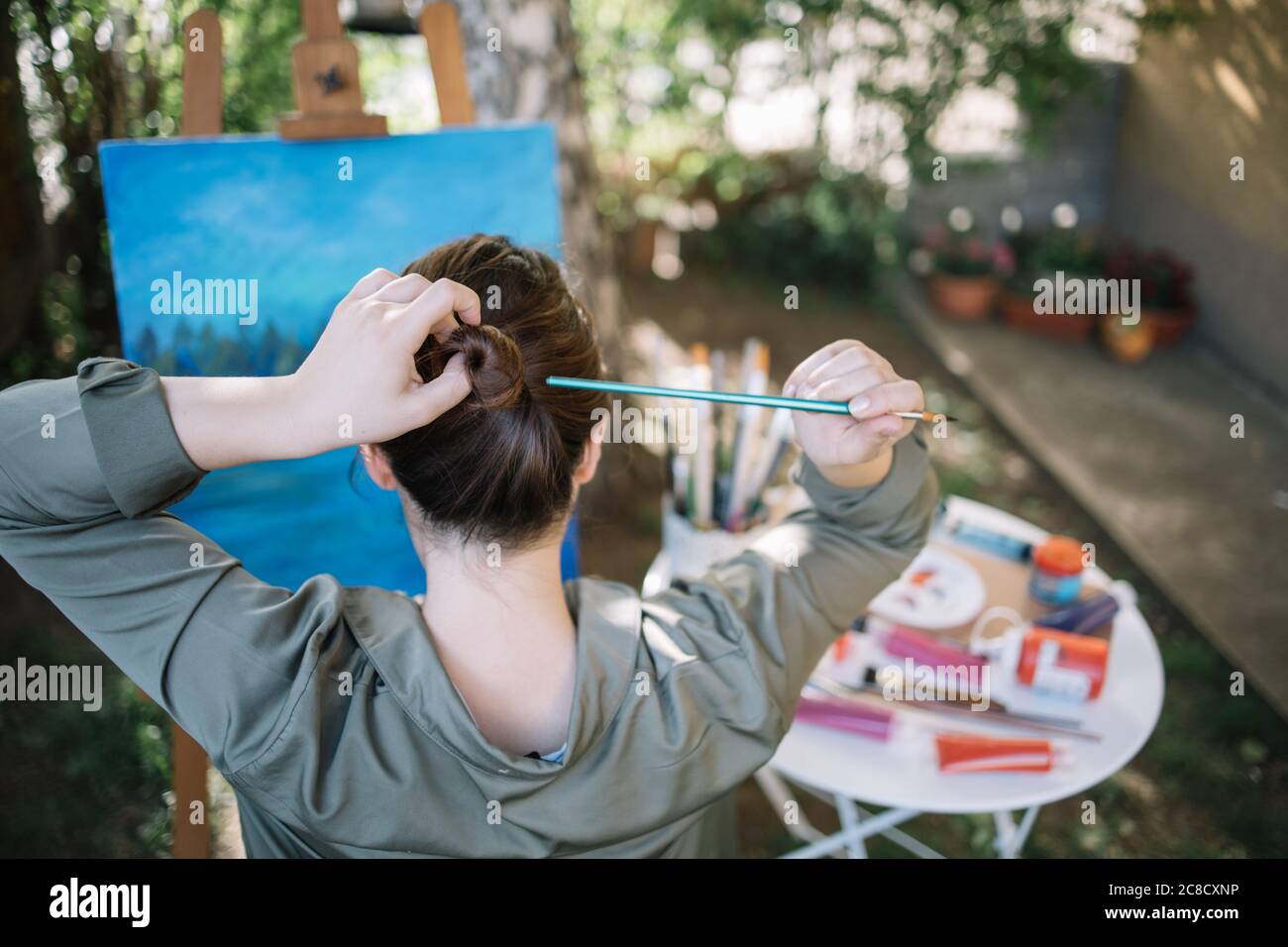 Girl making hair bun using paintbrush in front of painted canvas Stock