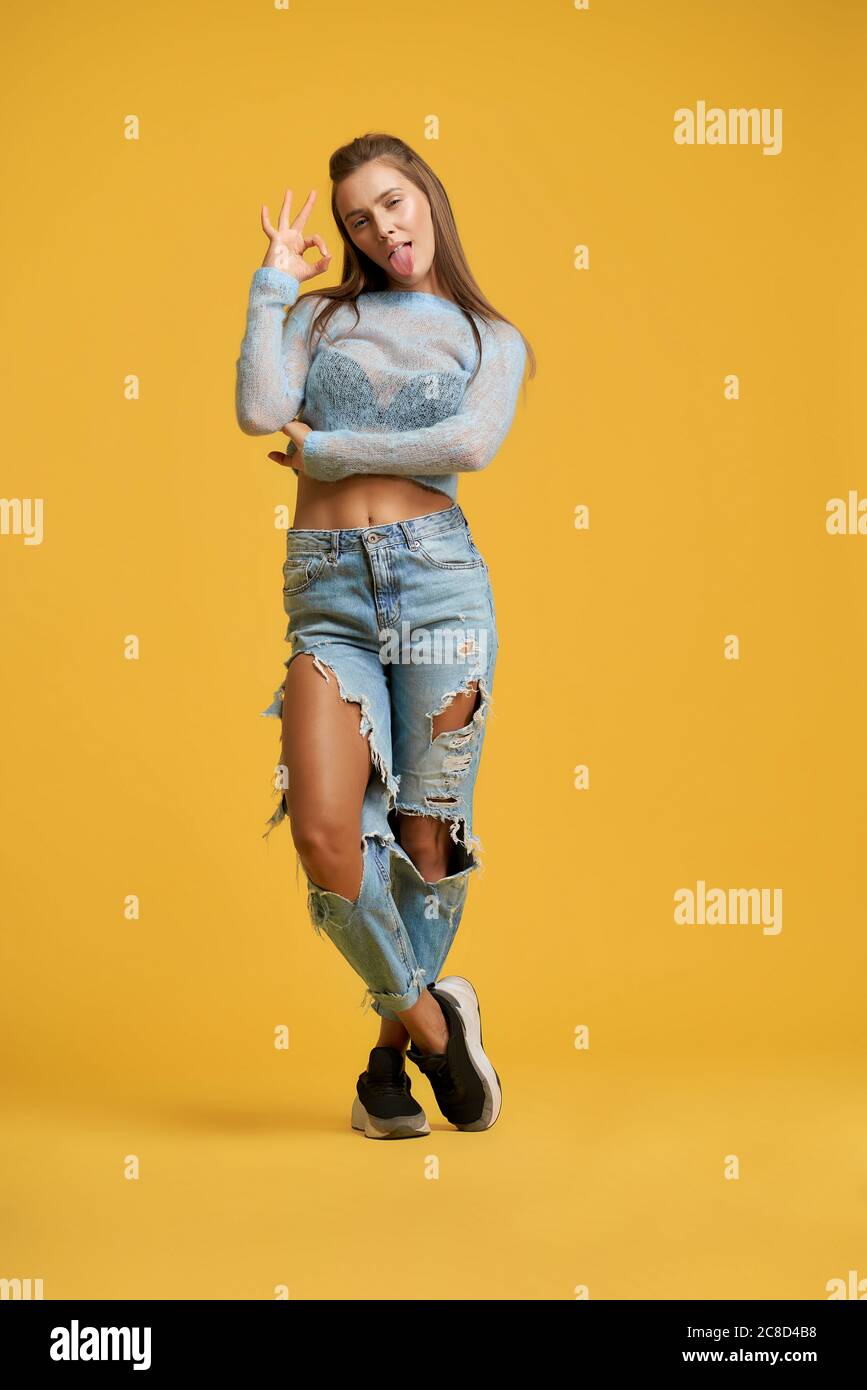 Front view of stylish beautiful girl sticking out tongue and doing ok symbol with hand. Pretty lady wearing ripped jeans and sweater. Isolated on yellow studio background. Stock Photo