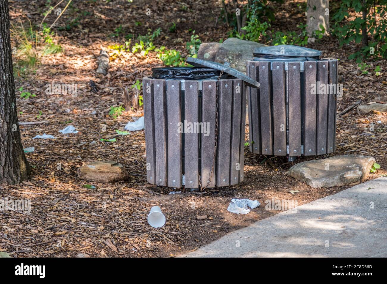 Garbage laying on the ground around two trash cans along the trail at the park by the woodlands polluting the environment on a sunny day in summertime Stock Photo