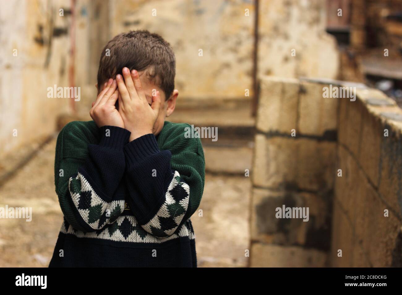 a poor Syrian child in slum covering his face with his hands, sneak peaking in between Stock Photo