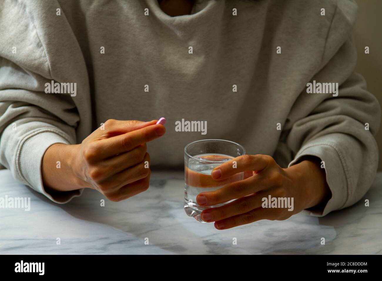 A woman is seen as she is holding a pill in one hand and a glass of water on the other hand. She is about to take the medication with a sip of water. Stock Photo