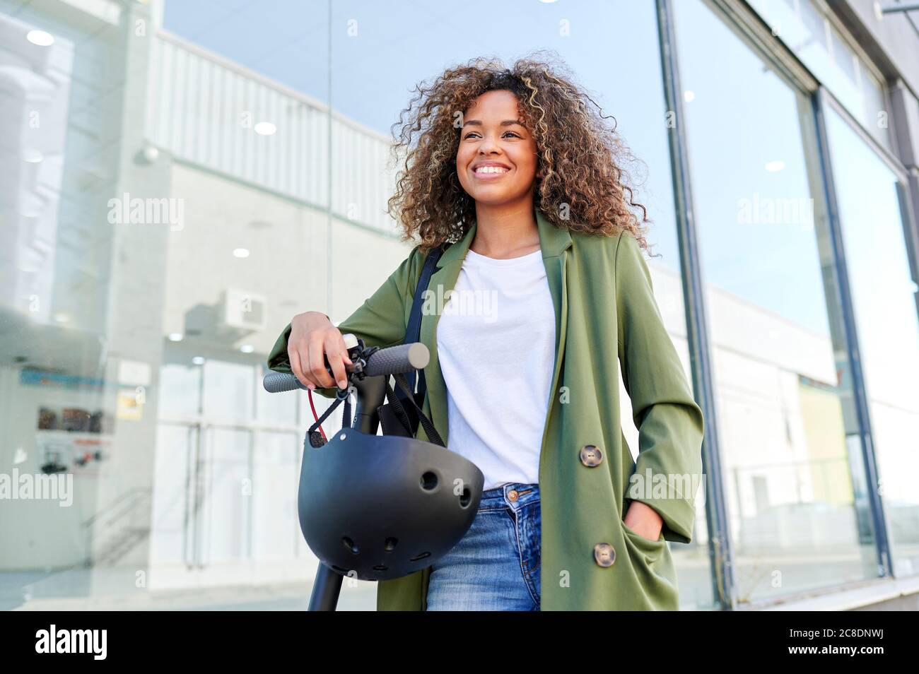 Thoughtful young woman smiling while standing with electric push scooter in city Stock Photo