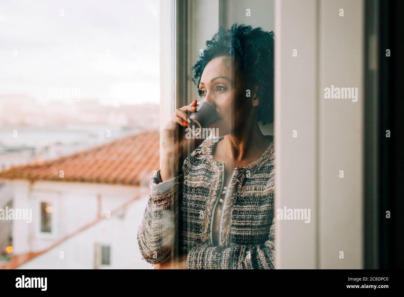 Woman drinking coffee while looking through window at home seen through glass door Stock Photo