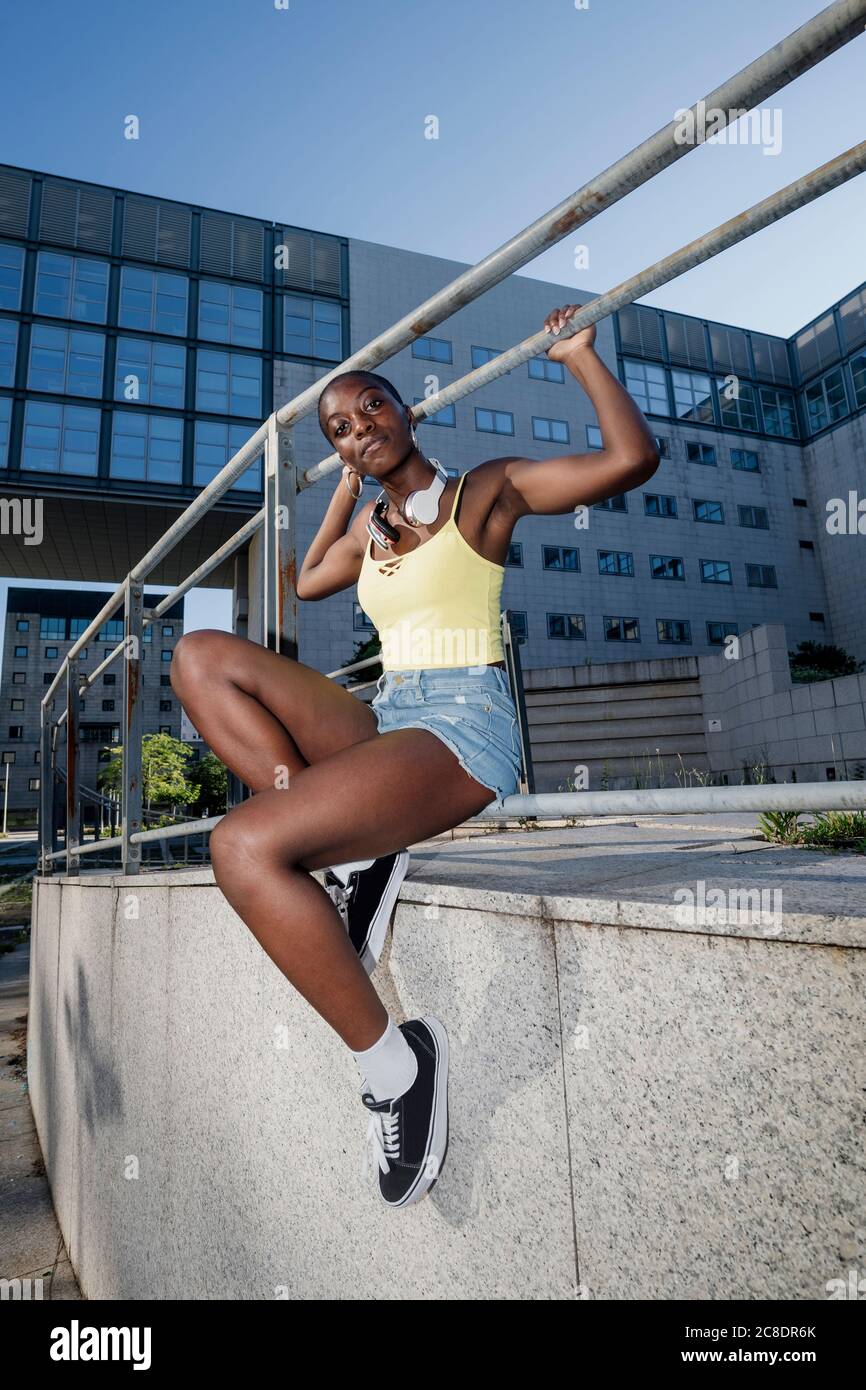 Young woman with shaved head sitting on railing against building in city Stock Photo