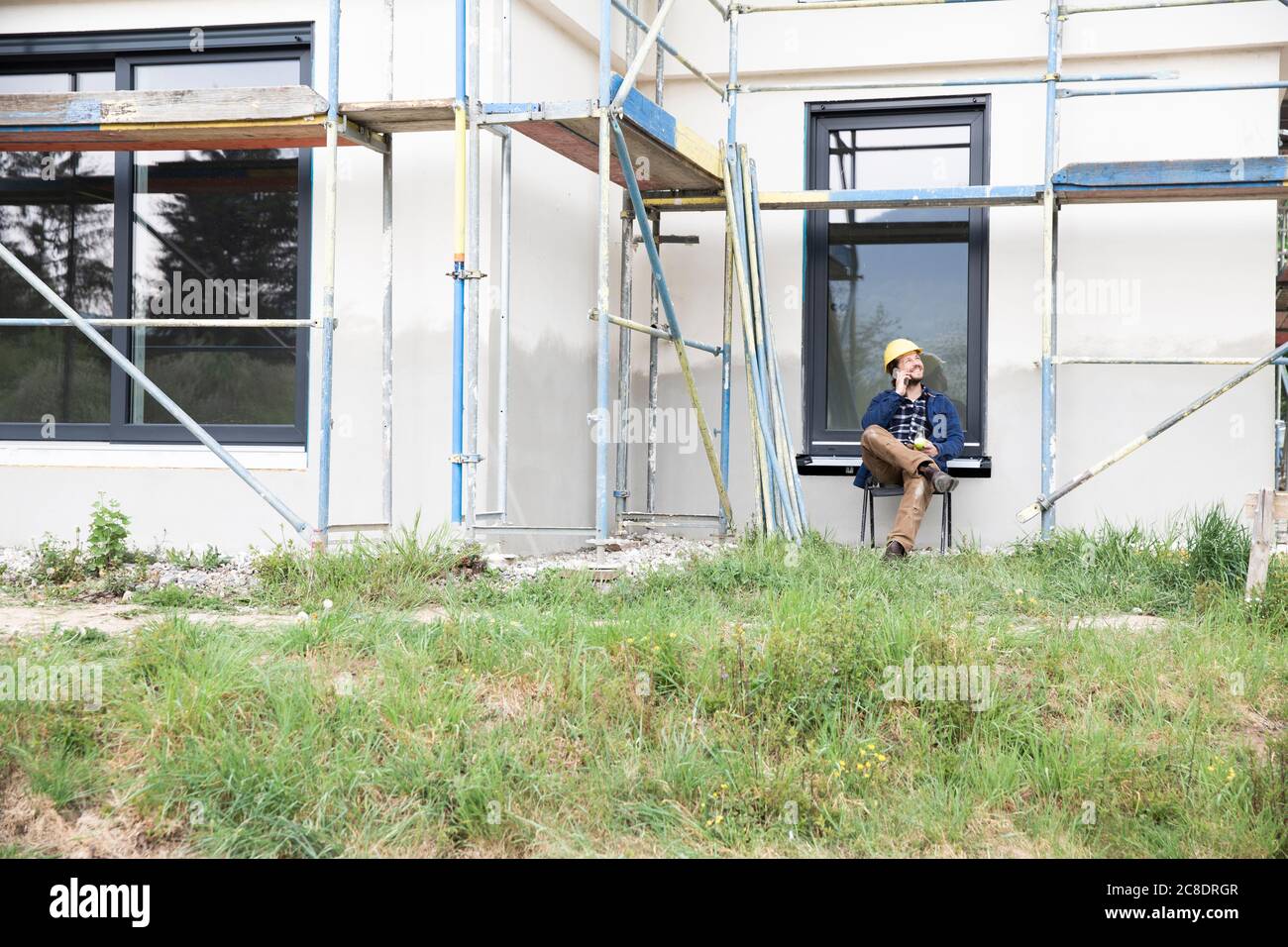 Construction worker talking over mobile phone while sitting against house Stock Photo