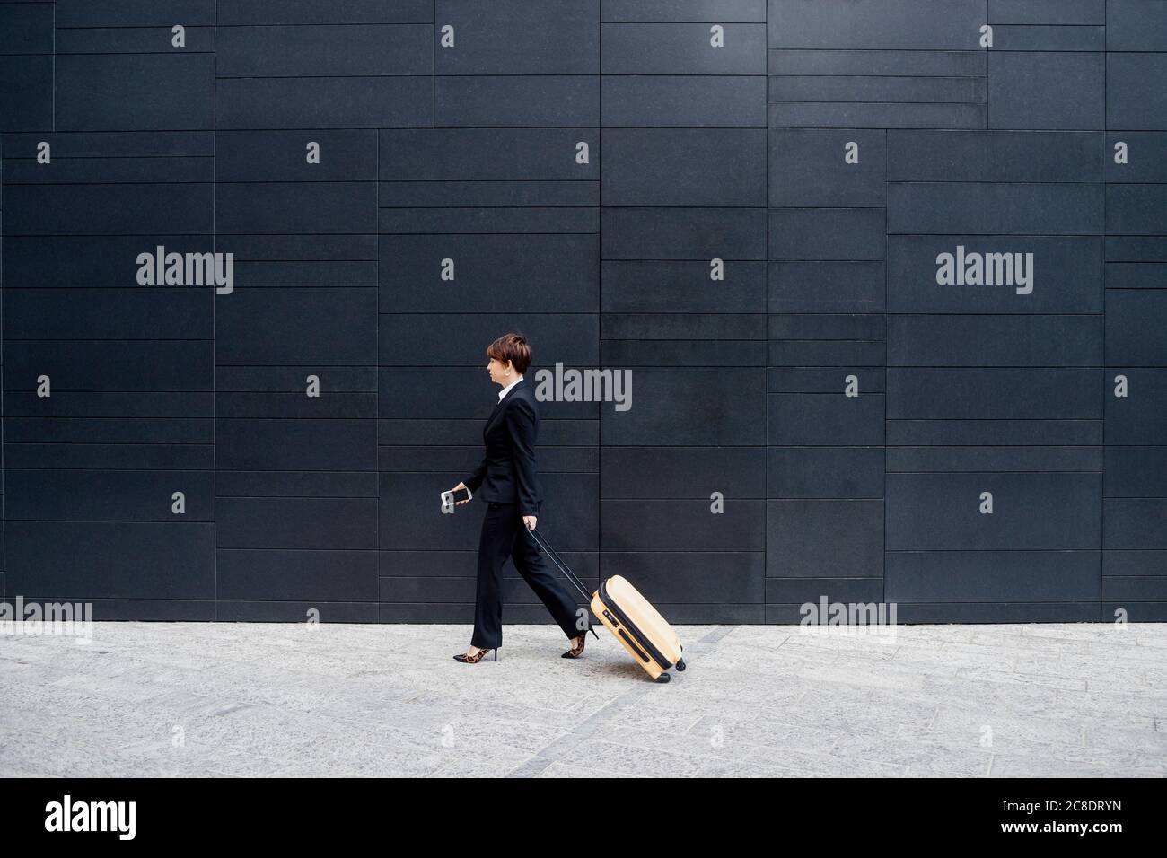 Businesswoman with suitcase walking on sidewalk by modern building in city Stock Photo
