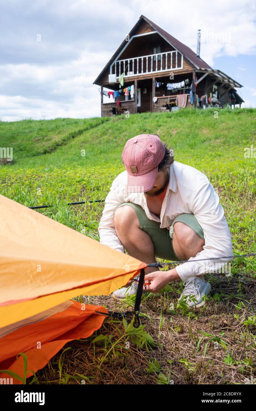 Mid adult man wearing cap installing tent on grassy land at campsite against sky Stock Photo