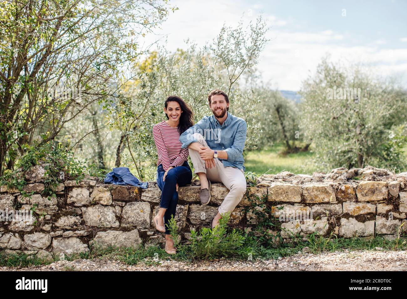 Smiling young couple sitting on retaining wall against trees Stock Photo