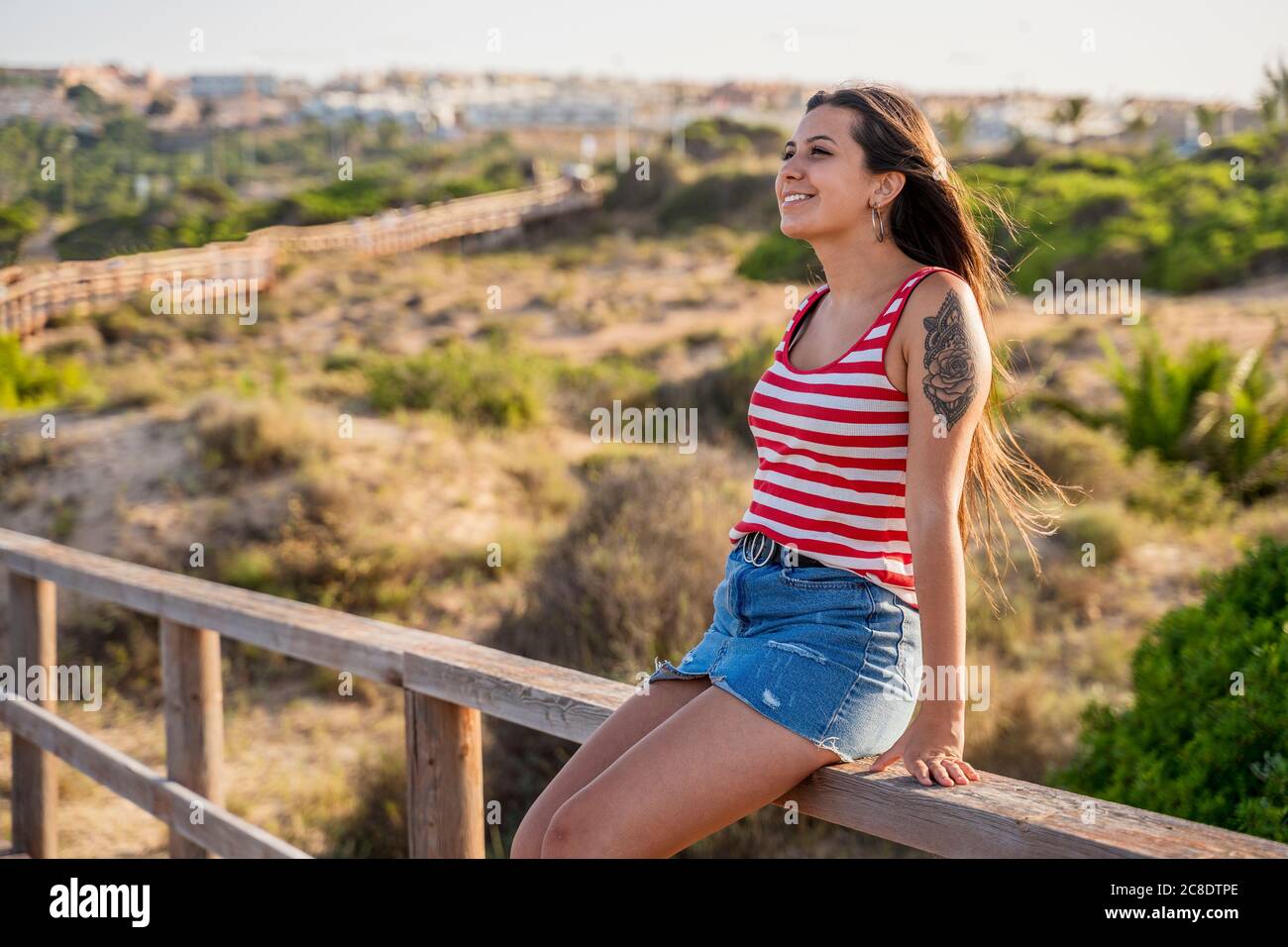 Thoughtful teenage girl sitting on wooden railing at beach during sunny day Stock Photo