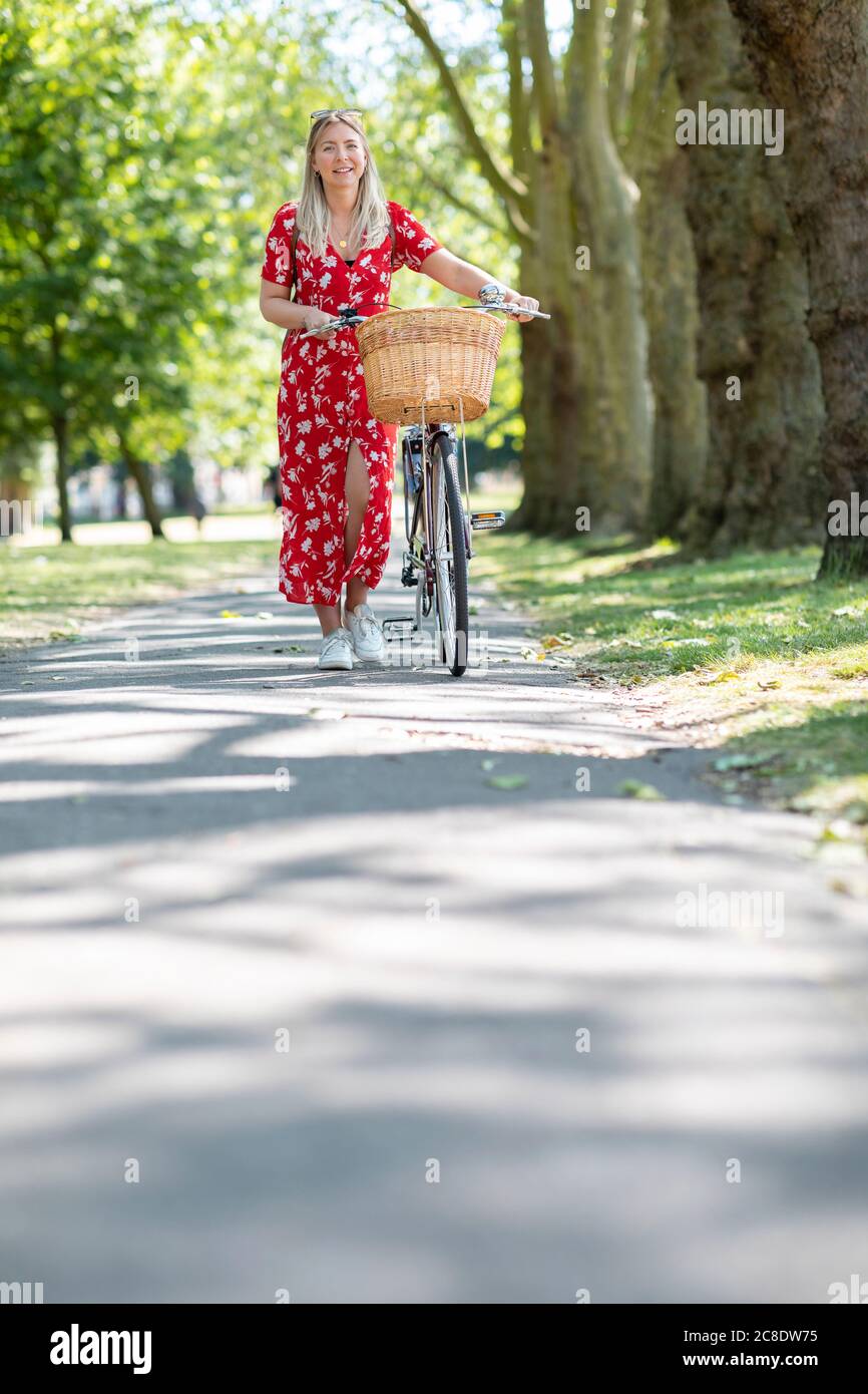 Happy woman walking with bicycle on footpath in public park during sunny day Stock Photo
