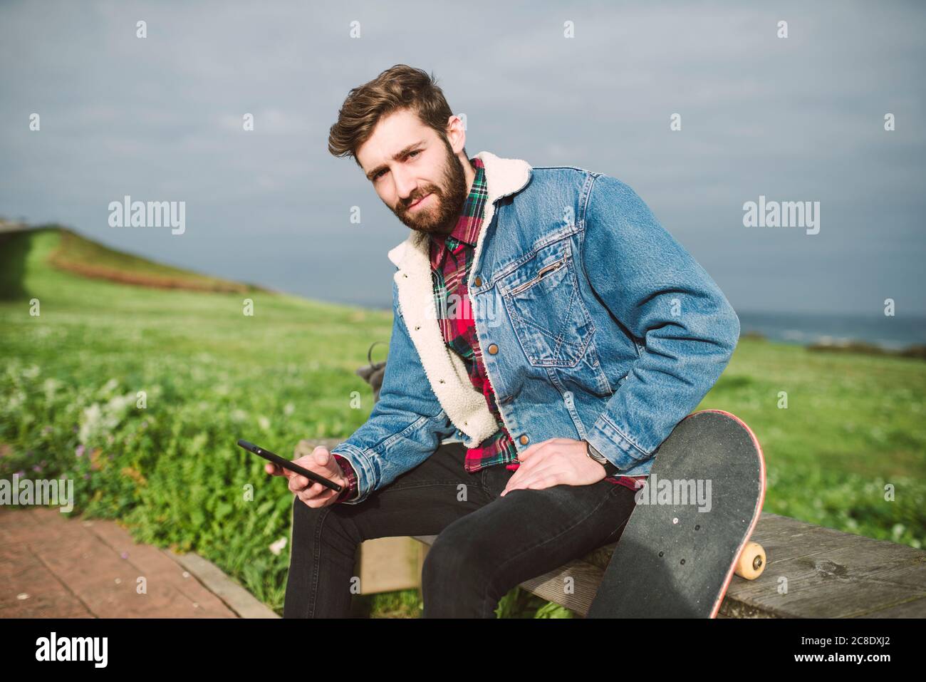 Handsome man using smart phone while sitting with skateboard on seat against cloudy sky Stock Photo