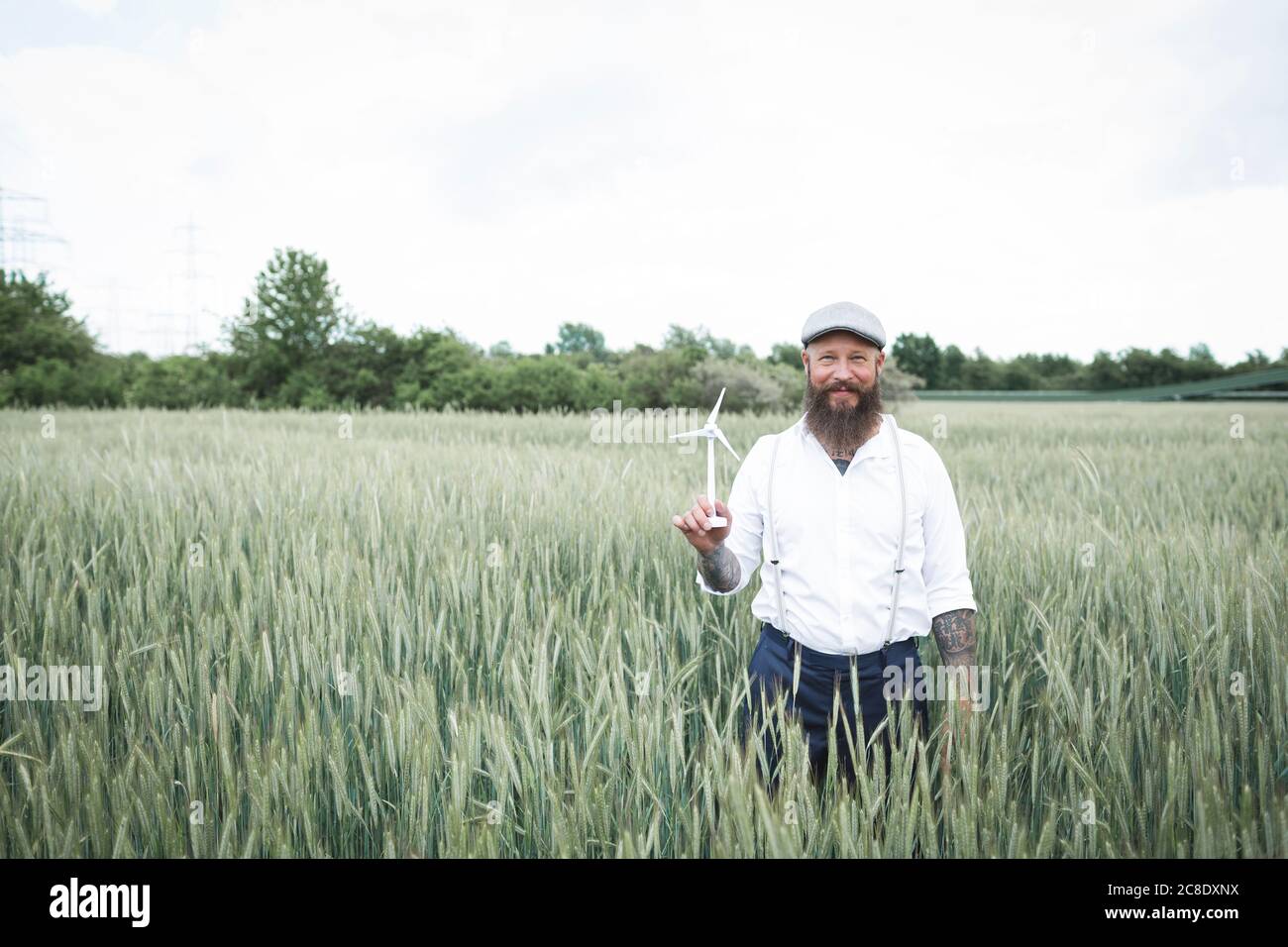 Bearded man holding small windmill while standing amidst cornfield against sky Stock Photo