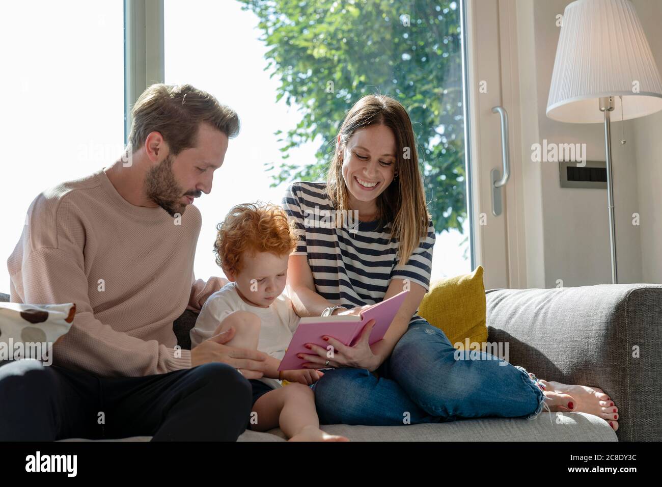 Smiling woman reading picture book while sitting by son and man on sofa in living room at home Stock Photo