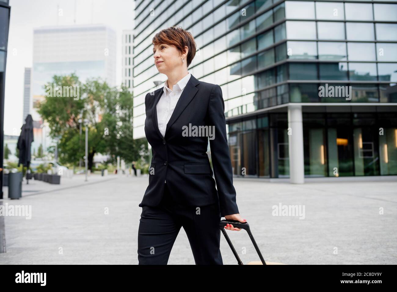Female entrepreneur with suitcase walking on street against modern building in city Stock Photo