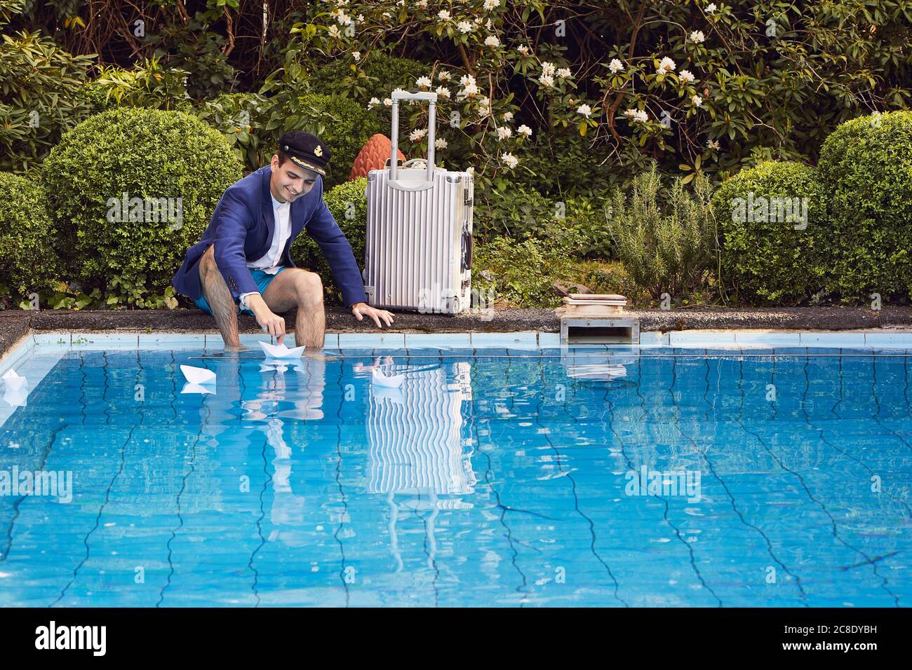 Man floating paper boats on swimming pool while sitting with suitcase against plants in yard Stock Photo