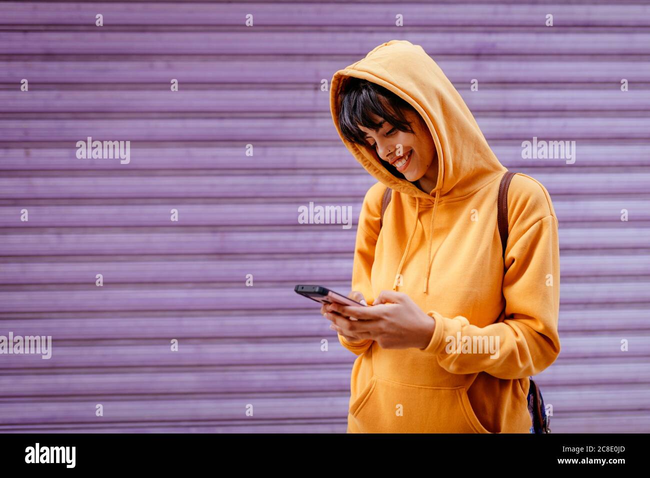 Portrait of young woman with yellow hoodie checking smartphone in front of purple background Stock Photo