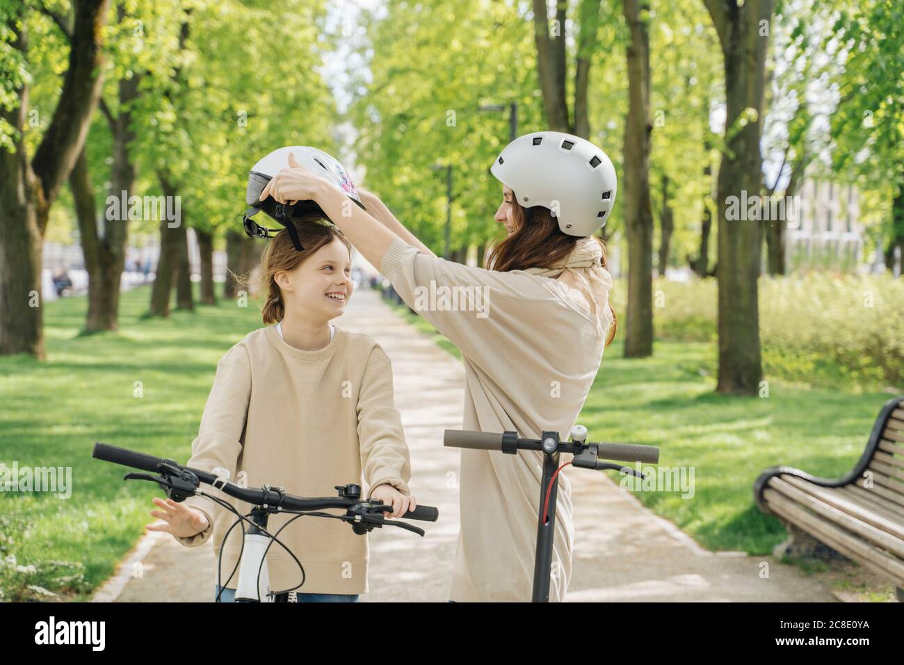 Mother wearing helmet to her daughter while standing against trees in park Stock Photo