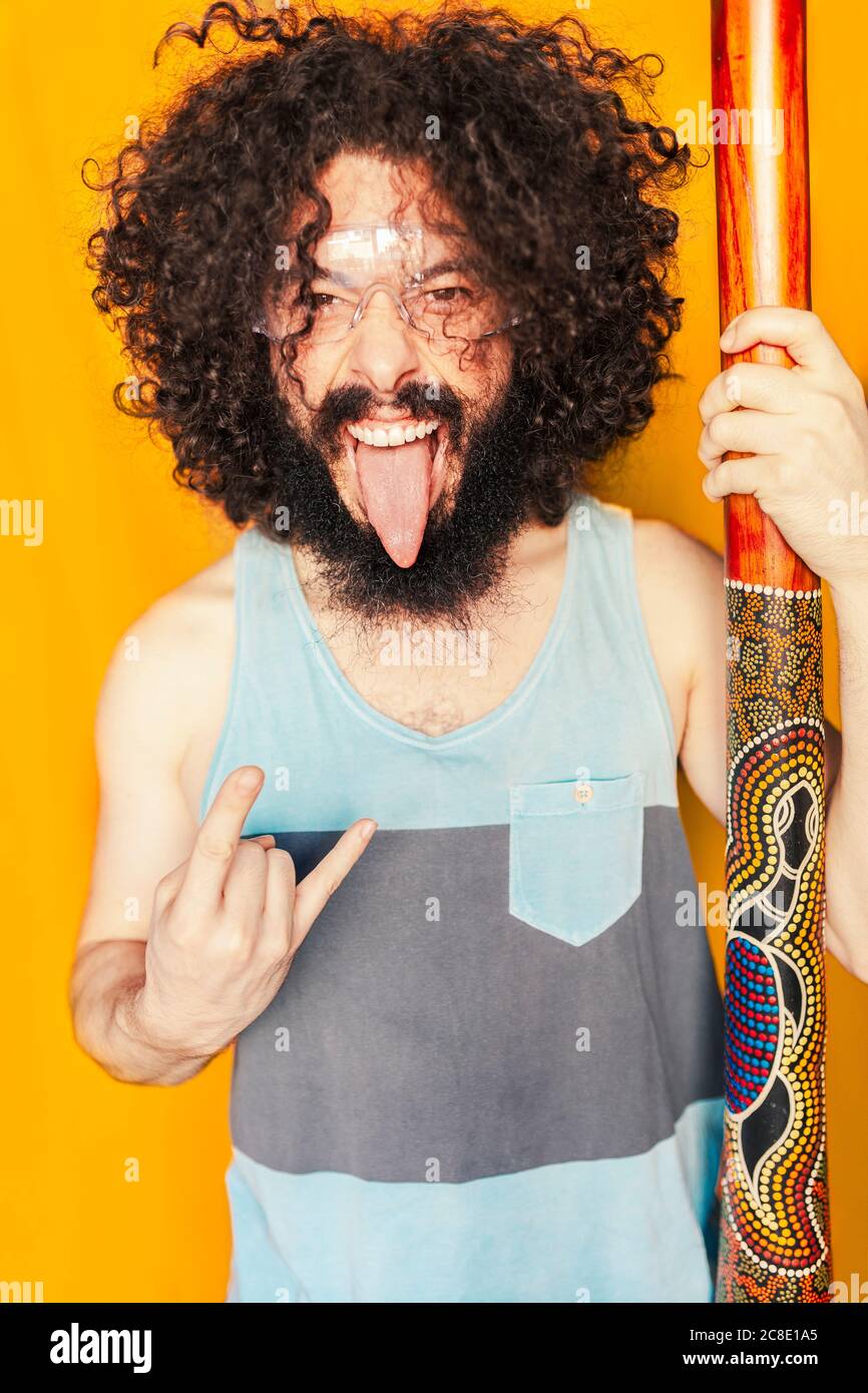 Crazy man with curly hair holding didgeridoo while sticking out tongue against yellow background Stock Photo
