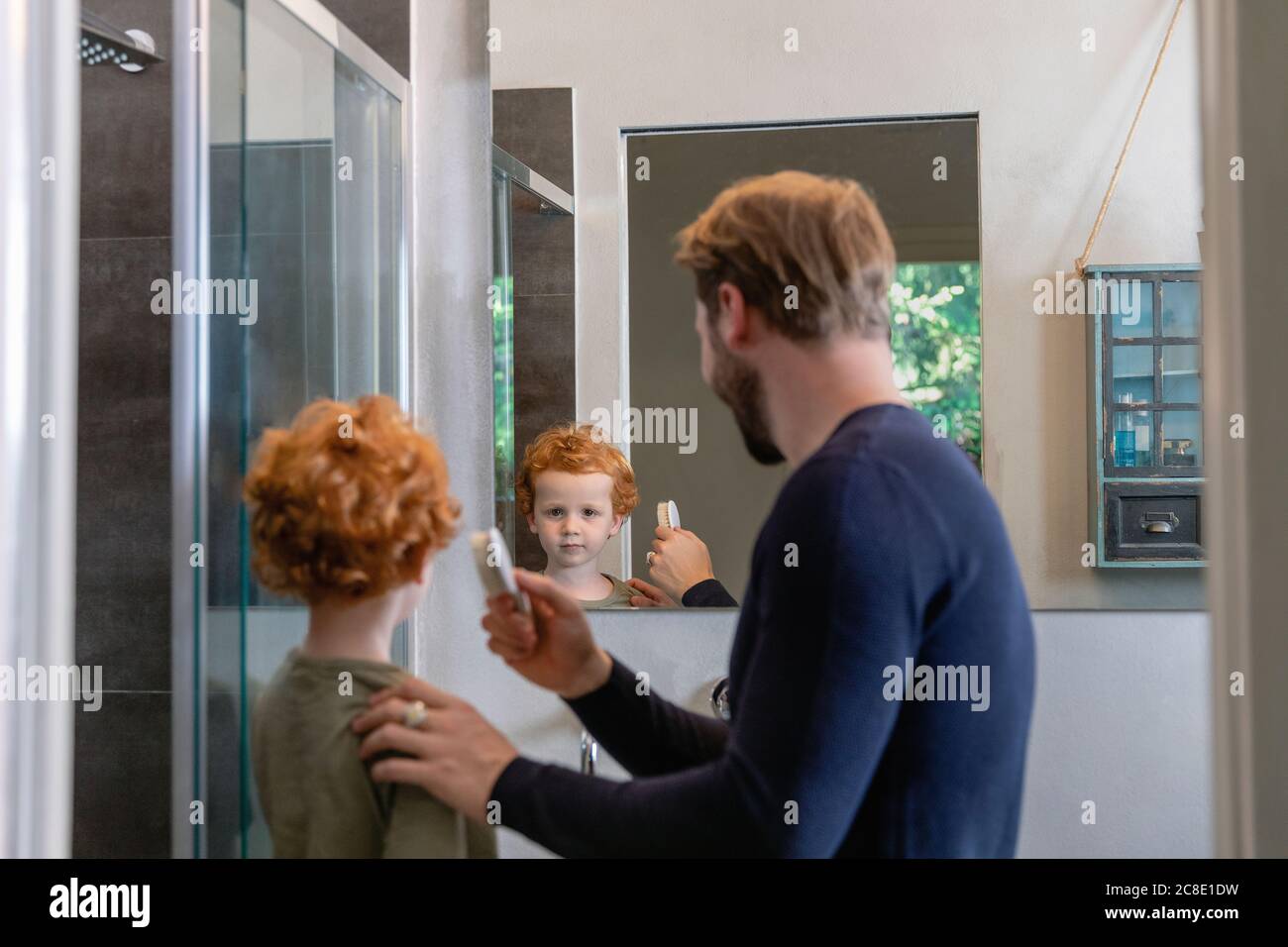 Cute boy looking at mirror reflection of father brushing his hair in