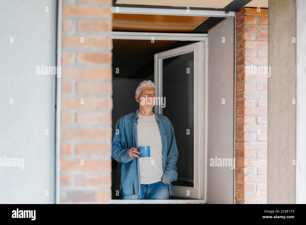 Thoughtful senior man holding coffee mug while looking through window Stock Photo