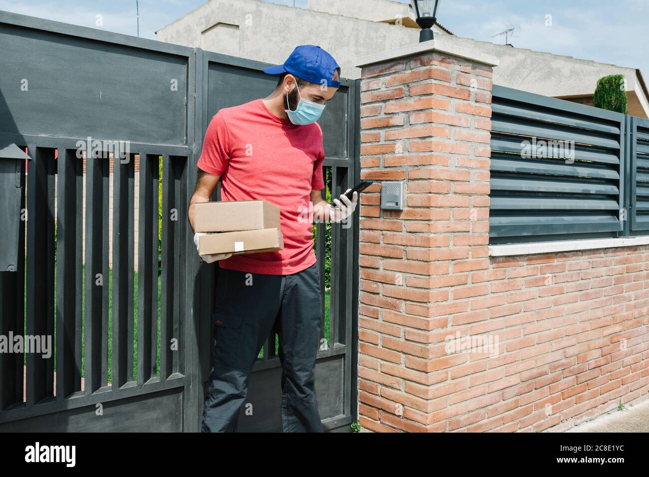 Male postal worker using mobile phone while standing with package against gate Stock Photo