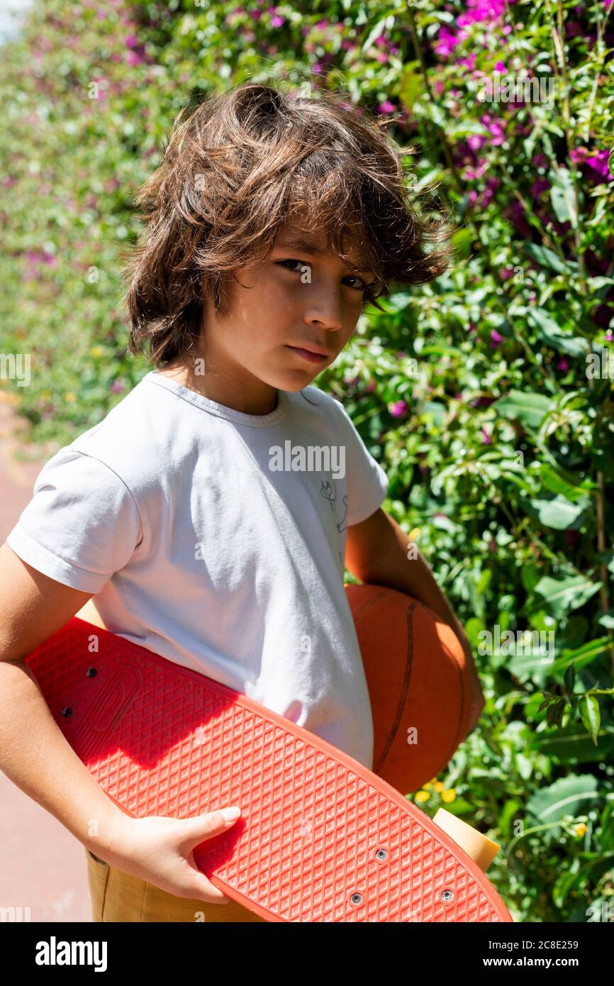 Close-up of boy with basketball and skateboard standing by plants in park Stock Photo