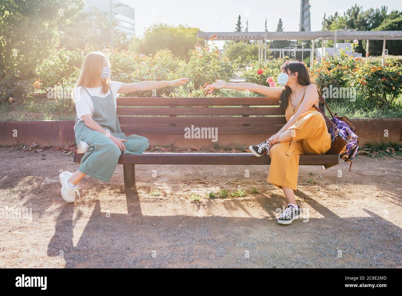 Two friends wearing protective masks sitting on bench during Corona crisis and keeping distance Stock Photo