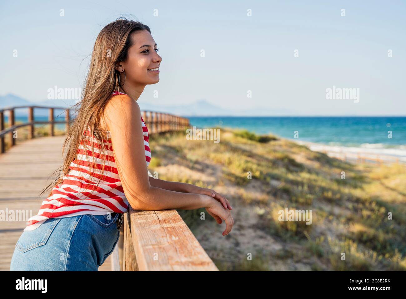 Thoughtful teenage girl standing on boardwalk at beach against clear blue sky Stock Photo